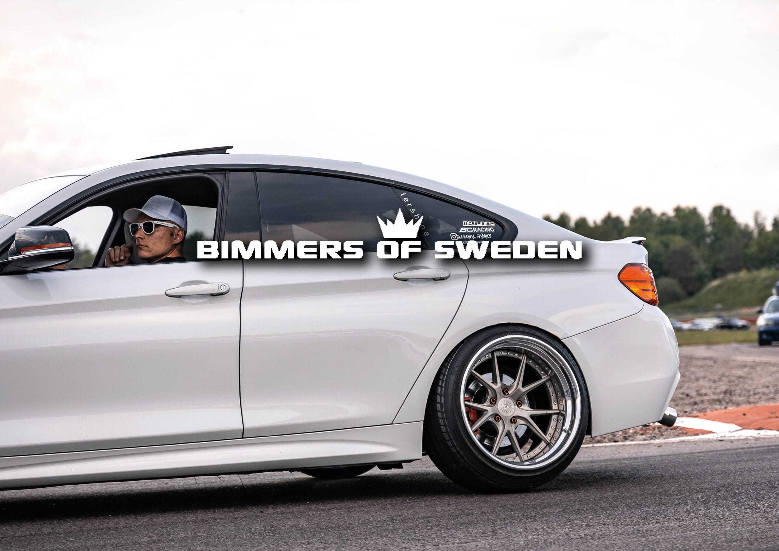 En person med solglasögon och keps kör en vit BMW sedan på en racerbana. Texten BIMMERS OF SWEDEN med en kronlogotyp visas över bilden. Träd och molnig himmel finns i bakgrunden.