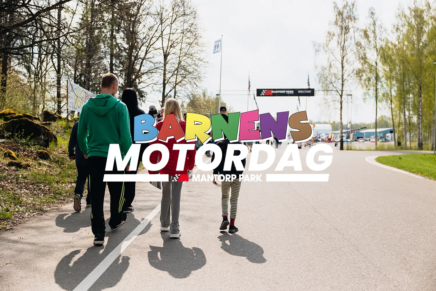 En grupp människor, inklusive barn, går längs en asfalterad stig mot en startlinje. Orden "Barnens Motordag Mantorp Park" står väl synligt över bilden. Glesa träd och grönska syns längs stigen.