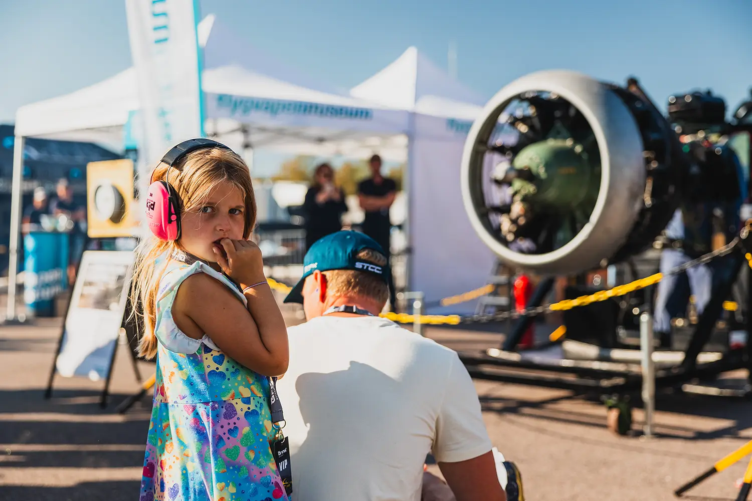 En ung tjej med blont hår klädd i rosa hörselkåpor och en färgglad klänning står bredvid en vuxen i vit skjorta på ett utomhusevenemang. Bakom dem står en jetmotor utställd, omgiven av gul tejp, med tält och en klarblå himmel i bakgrunden.