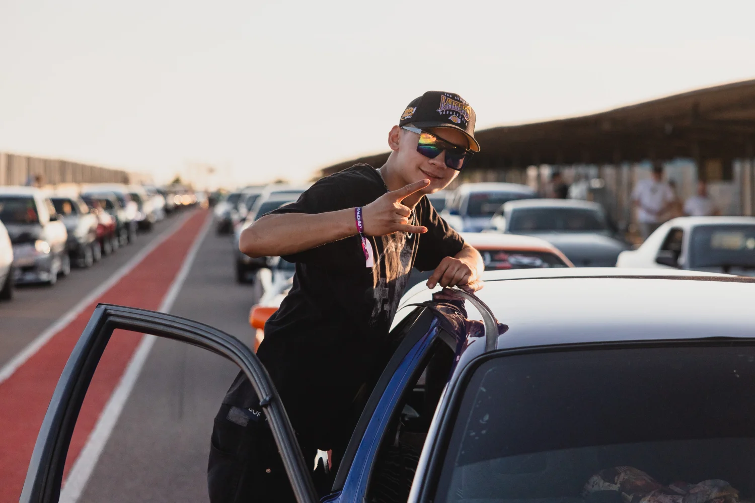 En person som bär solglasögon och keps lutar sig ut ur en blå bil på en väg kantad av parkerade bilar. De ler och gör ett fredstecken med fingrarna. Scenen är under en klar himmel.