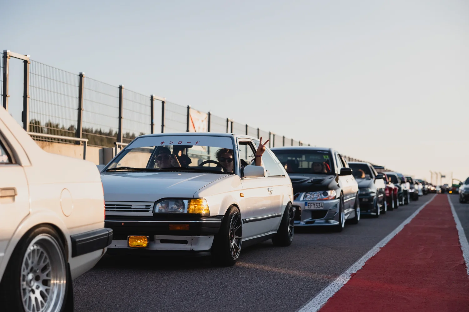 En rad snygga bilar, ledda av en vit sedan med en person inuti som gör en fredstecken, står parkerad på en solbelyst racerbana. En röd spårkant löper längs med, med ett kedjelänksstängsel och en klar himmel i bakgrunden.