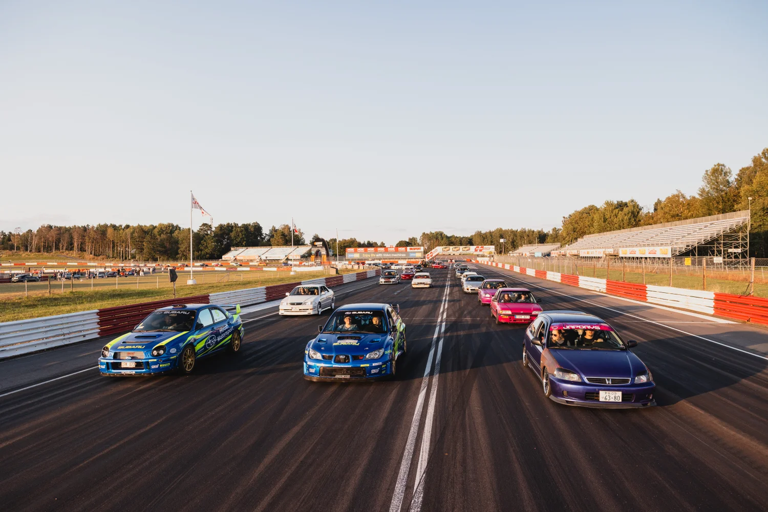 En grupp färgglada bilar, inklusive blå, grön, rosa och vita, ställde upp på en racerbana. Himlen är klar, och det finns träd och läktare i bakgrunden under solljus.