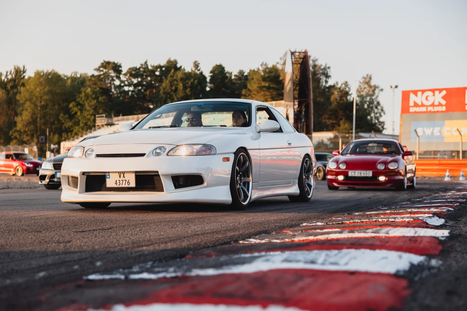 En vit, sportig bil leder en rad bilar på en racerbana, med solnedgången i bakgrunden. Andra bilar, inklusive en röd, följer noga. Banan är markerad med röd- och vitrandiga kanter. Träd och banderoller syns på avstånd.
