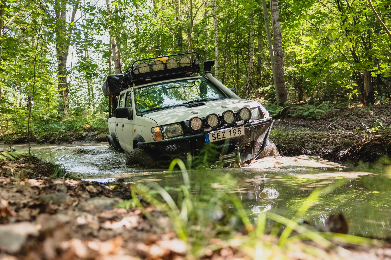 Ett vitt terrängfordon med takräcke och ljus kör genom en grund, lerig bäck i en tät skog. Solljus filtreras genom de gröna träden och vattnet stänker runt fordonet när det navigerar i den tuffa terrängen.