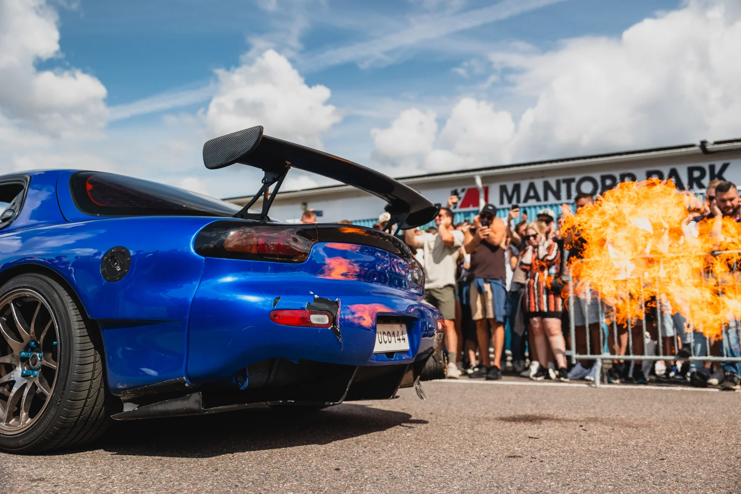 En blå sportbil med en stor bakvinge avger lågor från avgaserna. Åskådare står bakom ett räcke vid ett tävlingsevenemang under en delvis molnig himmel. En skylt i bakgrunden lyder "MANTORP PARK.