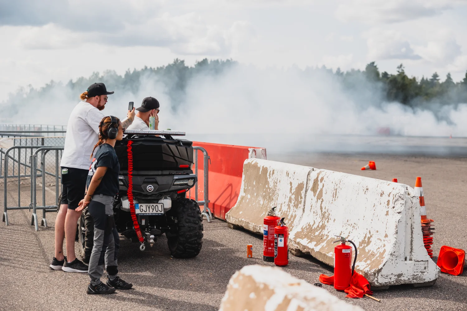Människor tittar på en rökig drifthändelse bakom säkerhetsbarriärer. De står bredvid ett svart fordon med bakluckan öppen. Brandsläckare placeras i närheten på marken. Bakgrunden visar ett skogsområde och molnig himmel.