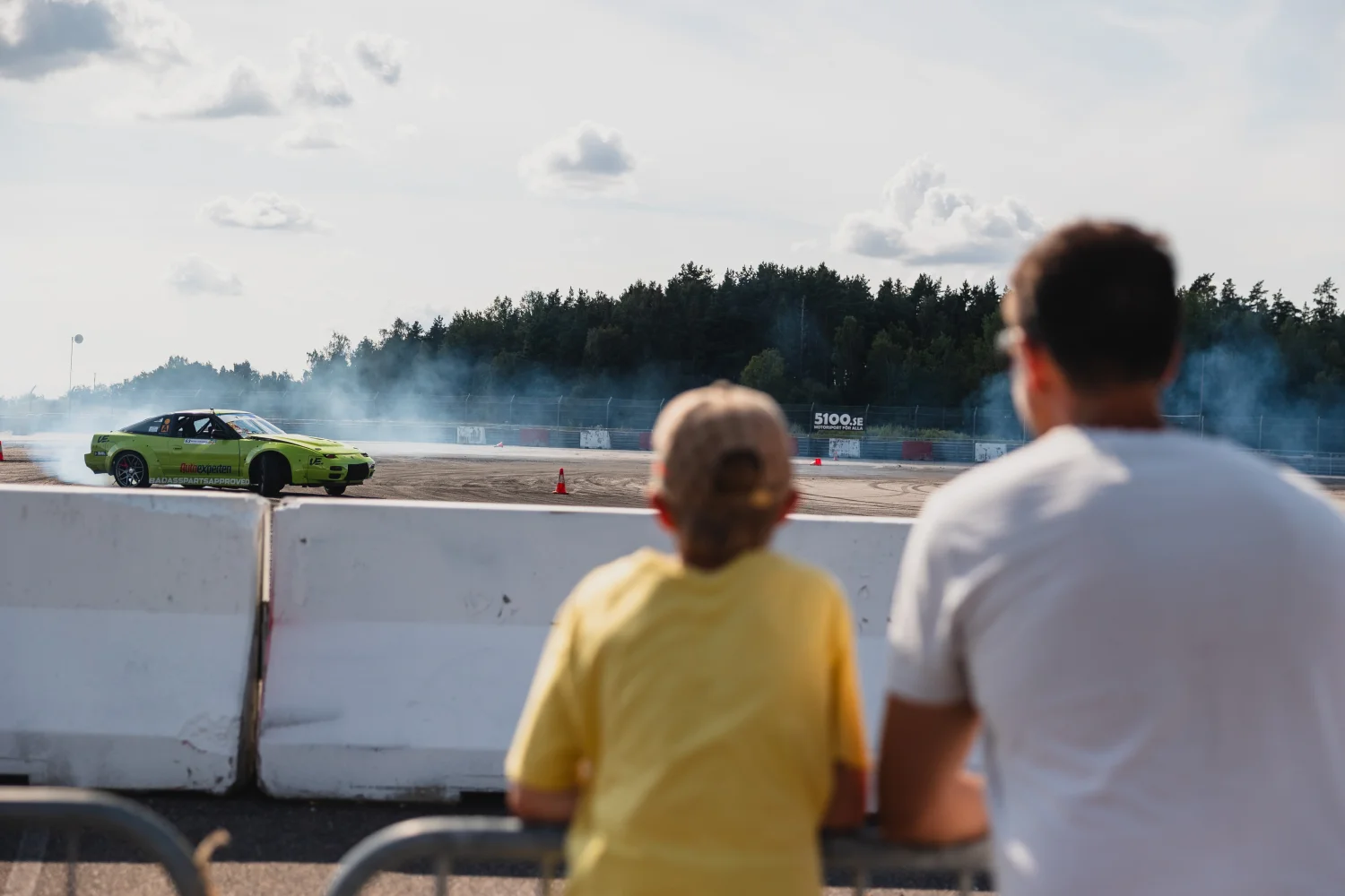 En gul sportbil driver på en racerbana och skapar rökspår. I förgrunden ser ett barn i gul skjorta och en vuxen bakom en barriär. Träd och en molnig himmel är i bakgrunden.