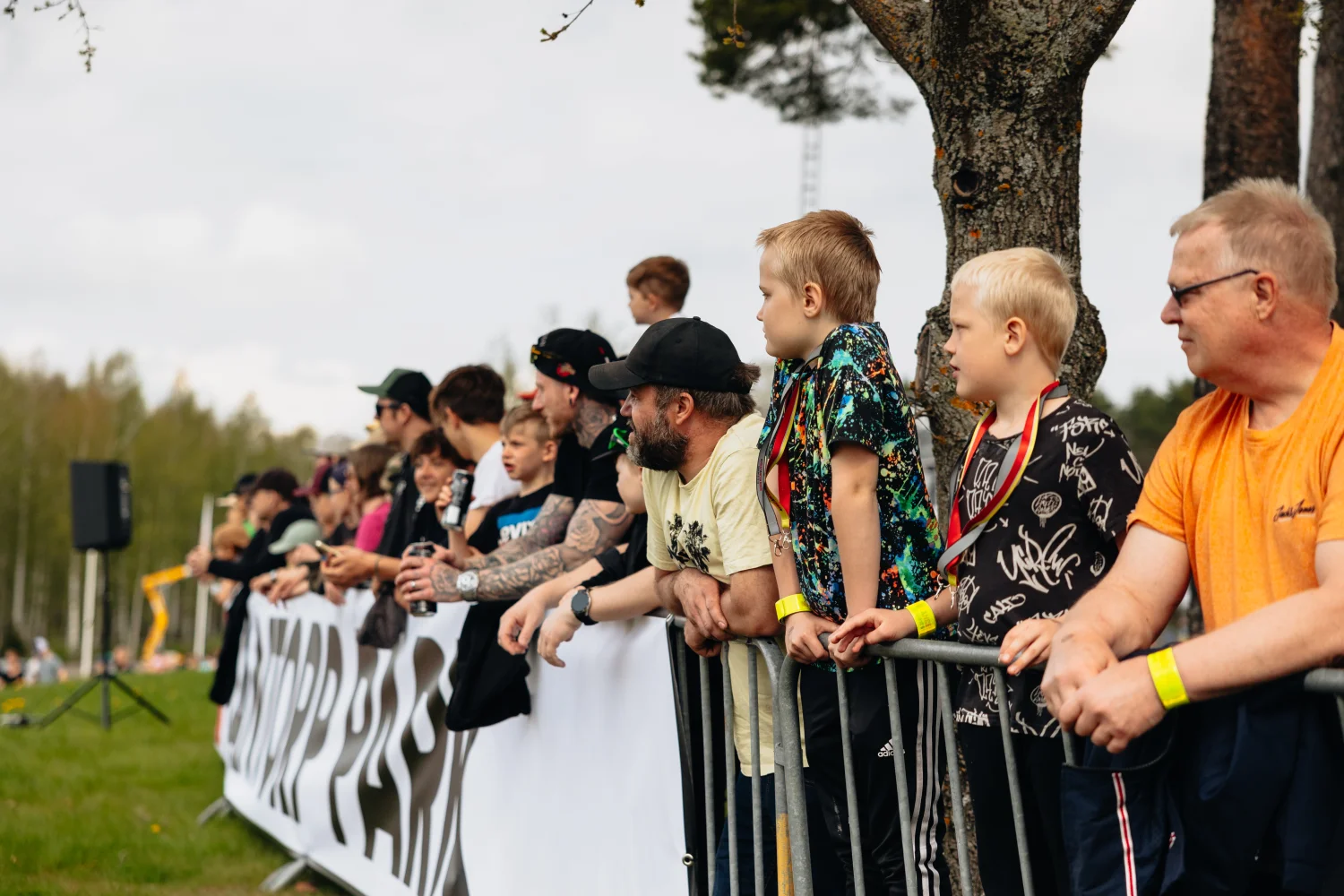 En grupp människor, inklusive vuxna och barn, står bakom en metallbarriär och tittar på ett utomhusevenemang. De har på sig vardagskläder och vissa har armband. Träd och en gräsyta syns i bakgrunden.