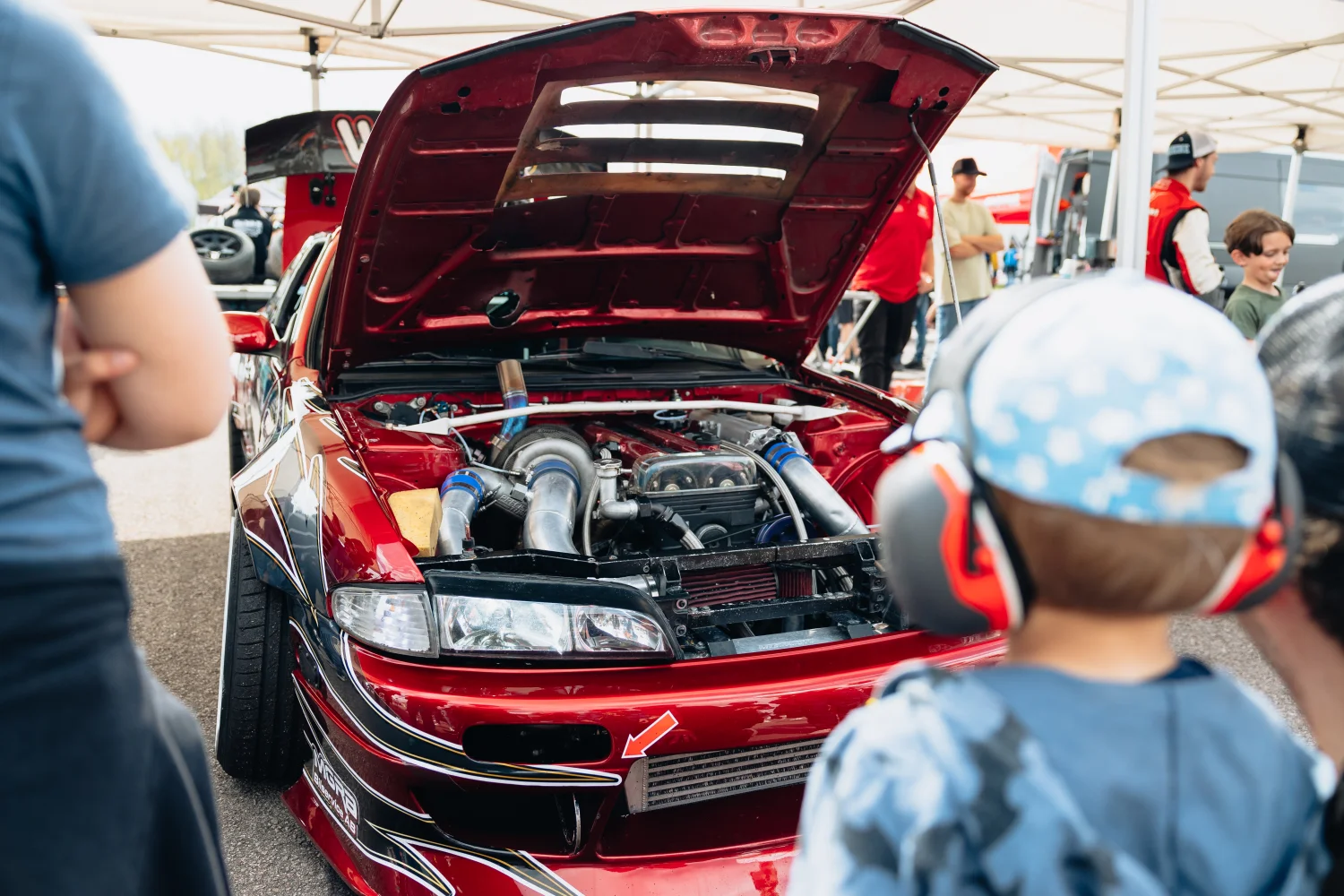 En knallröd modifierad bil med öppen huva visas. Flera personer, inklusive ett barn som bär hörlurar, observerar fordonet. Inramningen verkar vara ett bilevenemang utomhus med tält och en klar himmel i bakgrunden.