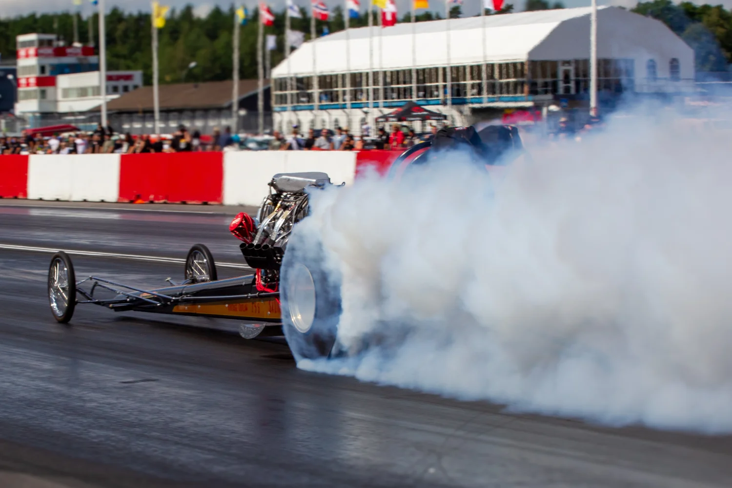 En dragracingbil rusar nerför en bana och producerar ett stort moln av vit rök från dess däck. Banan är kantad av röda och vita barriärer, och en folkmassa tittar på i bakgrunden mot en bakgrund av flaggor och byggnader.