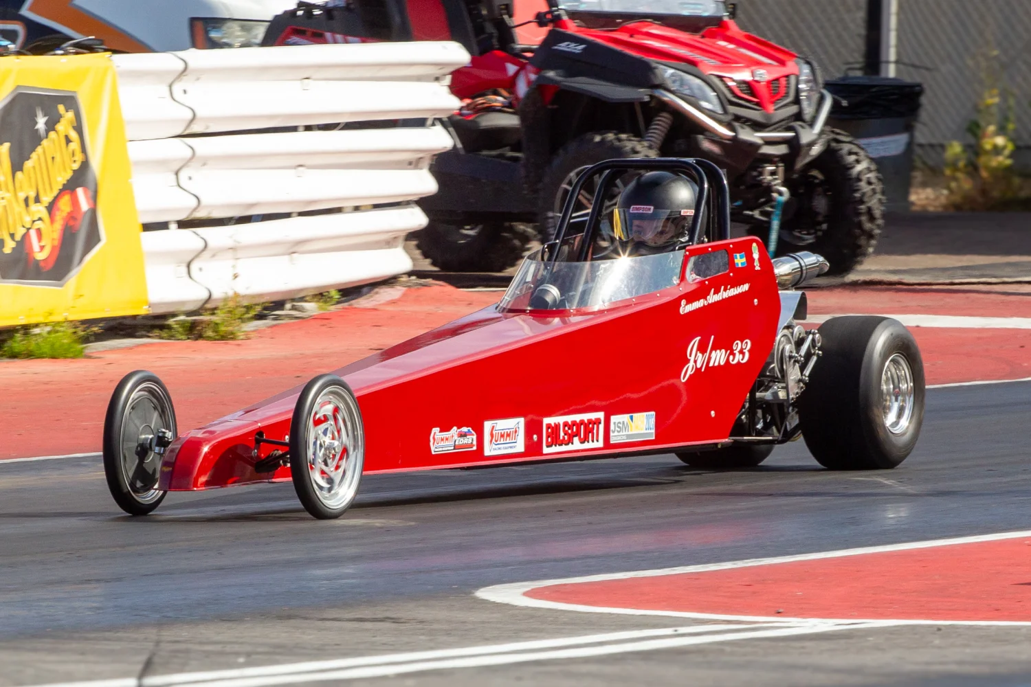 En röd dragracingbil rusar nerför banan med reklam på kroppen. Föraren är synlig och bommar kantar banan. Ett rött fordon och ett staket är i bakgrunden.