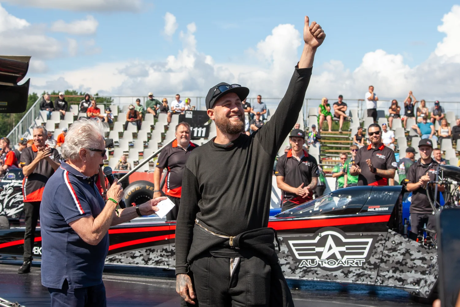 En person i svart outfit står på en racerbana och gör tummen upp, medan en annan person i blå skjorta håller i en mikrofon. En racerbil med svart och röd design står i bakgrunden, med åskådare som sitter på läktaren.