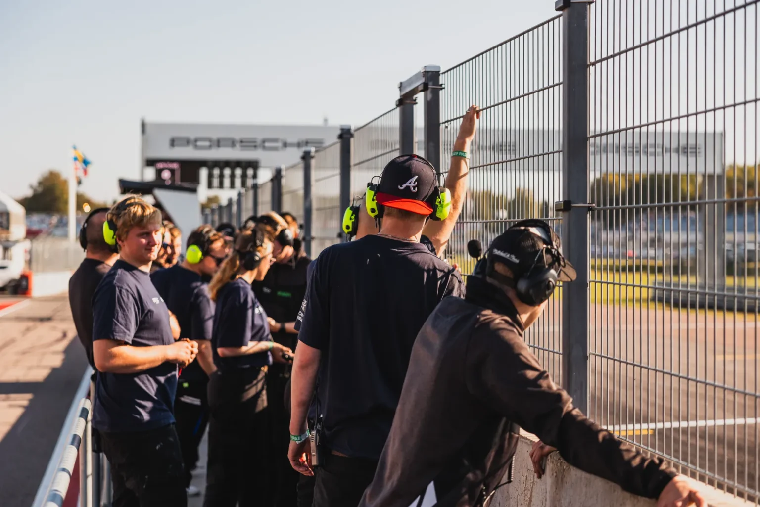 Människor som bär hörlurar står bredvid en racerbanas staket och tittar på en händelse. De är klädda i mörka kläder med säkerhetsutrustning. Bakgrunden visar ett spårområde med en "Porsche"-skylt och klar himmel.