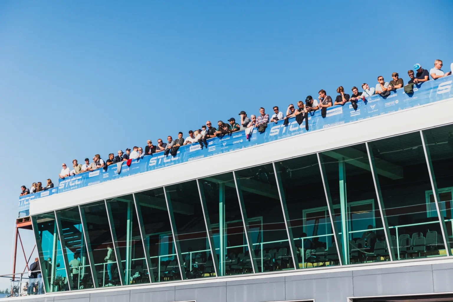 En stor skara människor står på en förhöjd plattform med ett blått räcke, med utsikt över en händelse. Plattformen ligger ovanpå en modern glasbyggnad. Himlen är klar och blå.
