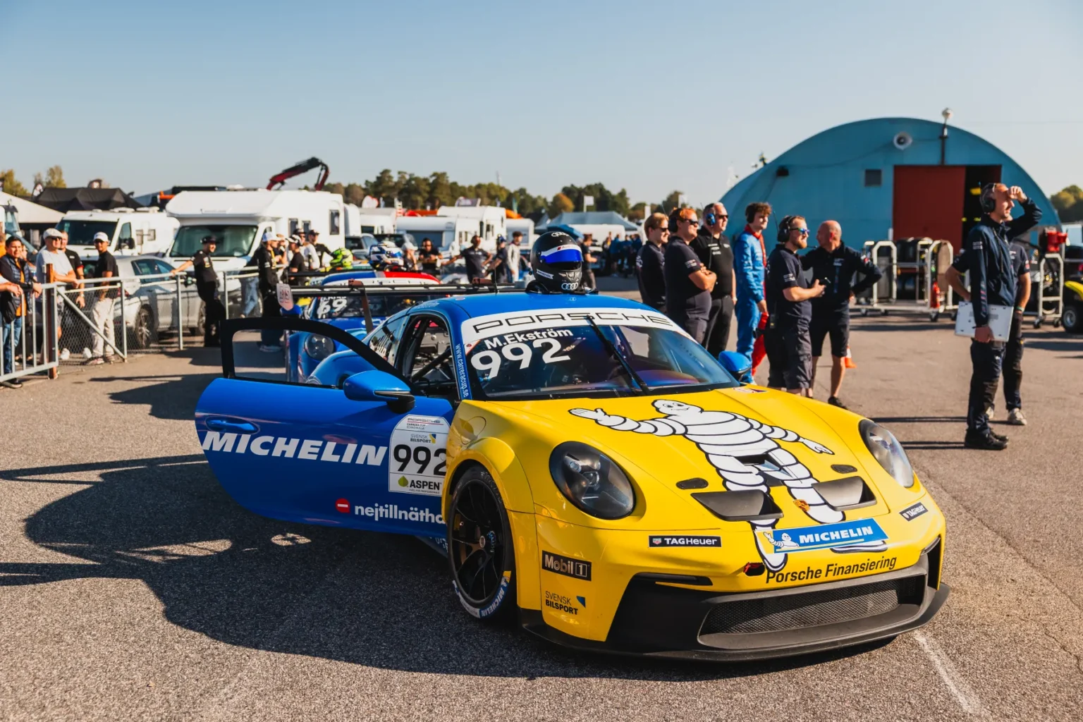 En gul och blå Porsche racerbil med nummer 992 står parkerad vid ett evenemang. Bilen har Michelin-märke. Människor i bakgrunden står nära släpvagnar och en välvd byggnad under en klar himmel.