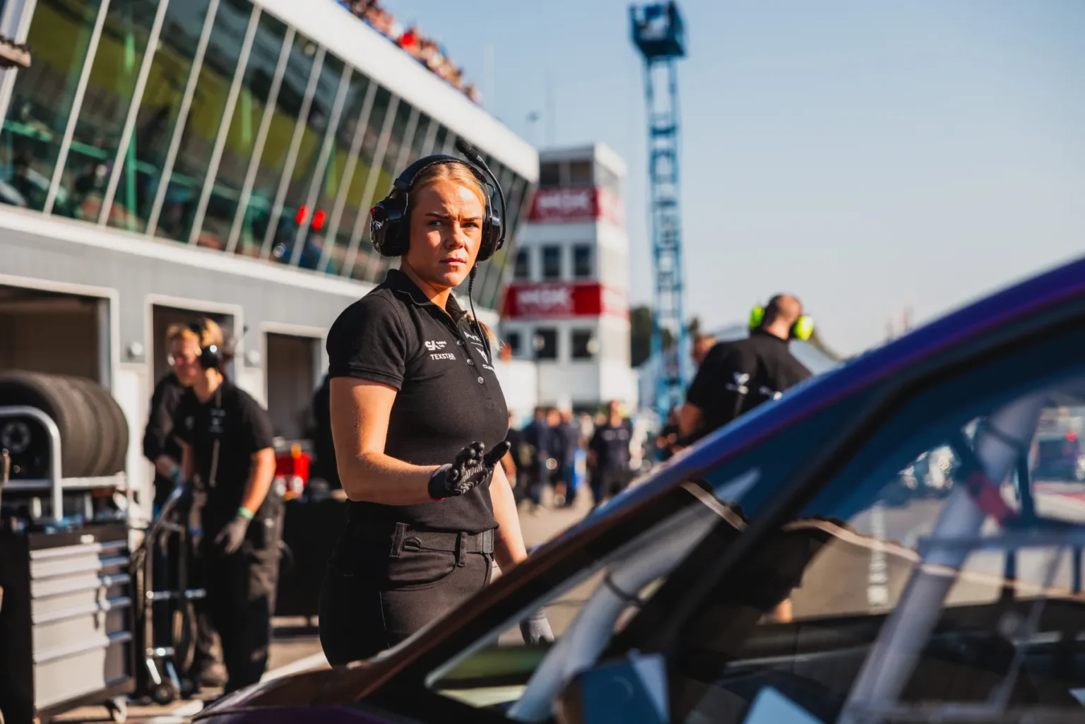 En kvinna i svart uniform och hörlurar står nära en racerbil på en upptagen racerbana. Hon verkar fokuserad, med teammedlemmar och utrustning i bakgrunden. Den soliga dagen framhäver den livliga, actionfyllda atmosfären.