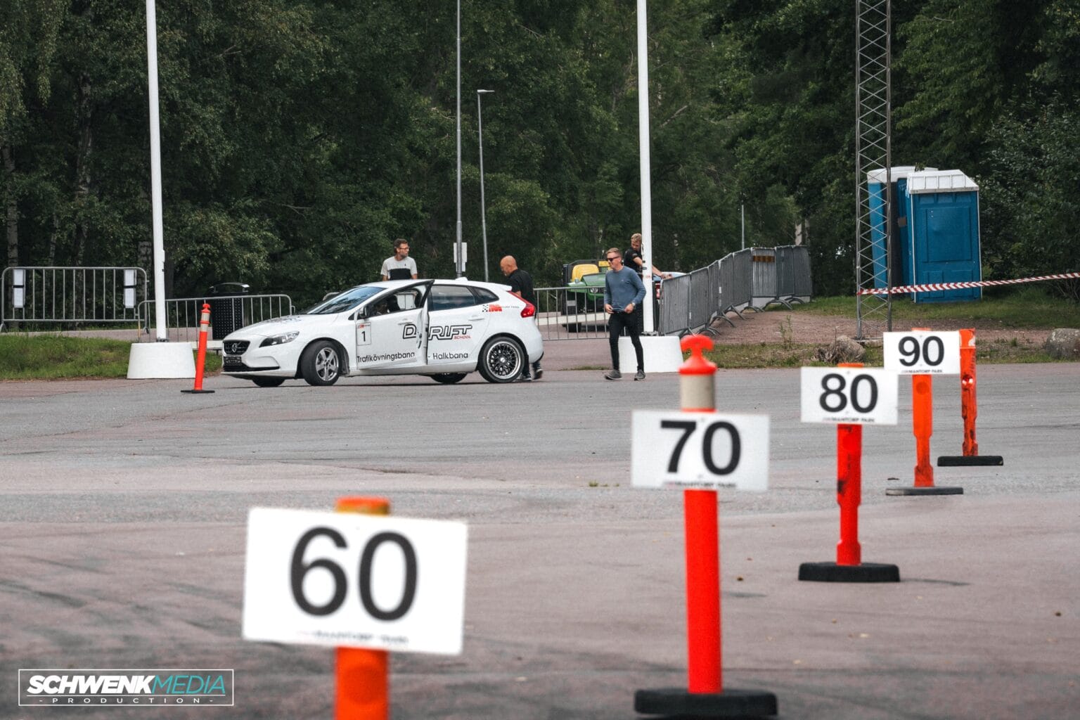 En vit bil står parkerad nära en körbana markerad med skyltar för hastighetsbegränsning: 60, 70, 80 och 90. Två personer står i närheten på en asfalterad yta. Det finns träd, en bärbar toalett och inhägnade områden i bakgrunden.
