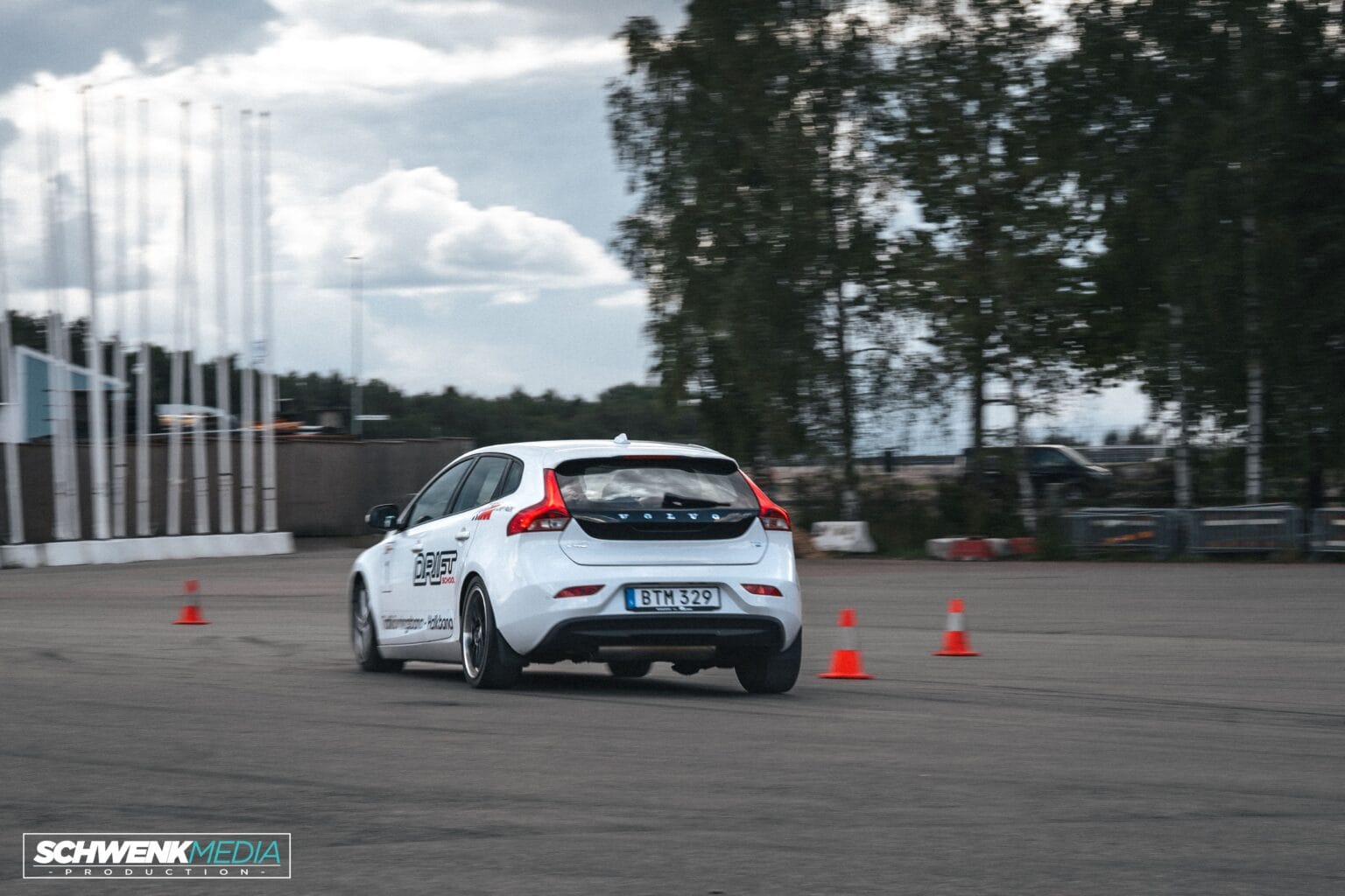 En vit halvkombibil manövrerar snabbt runt orangea kottar i ett öppet asfaltområde, troligen deltar i ett körevenemang. Träd och en molnig himmel syns i bakgrunden.
