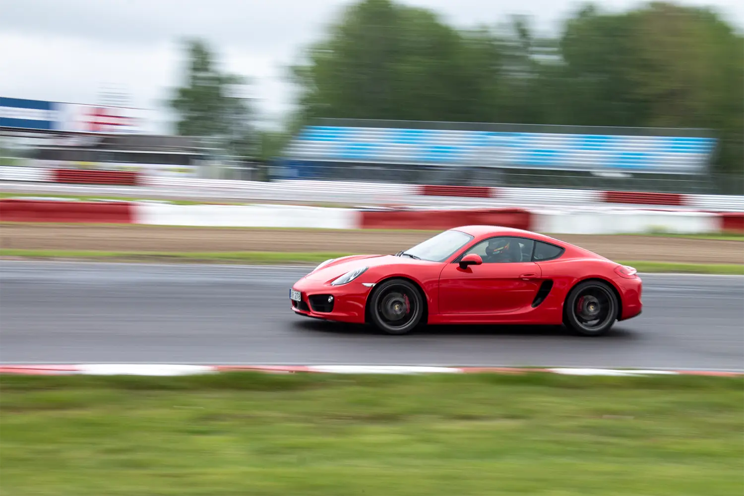 En röd sportbil rusar nerför en bana, med en rörelseoskärpa effekt som förmedlar hög hastighet. Bakgrunden har suddiga element på racerbanan och avlägsna träd under en molnig himmel.