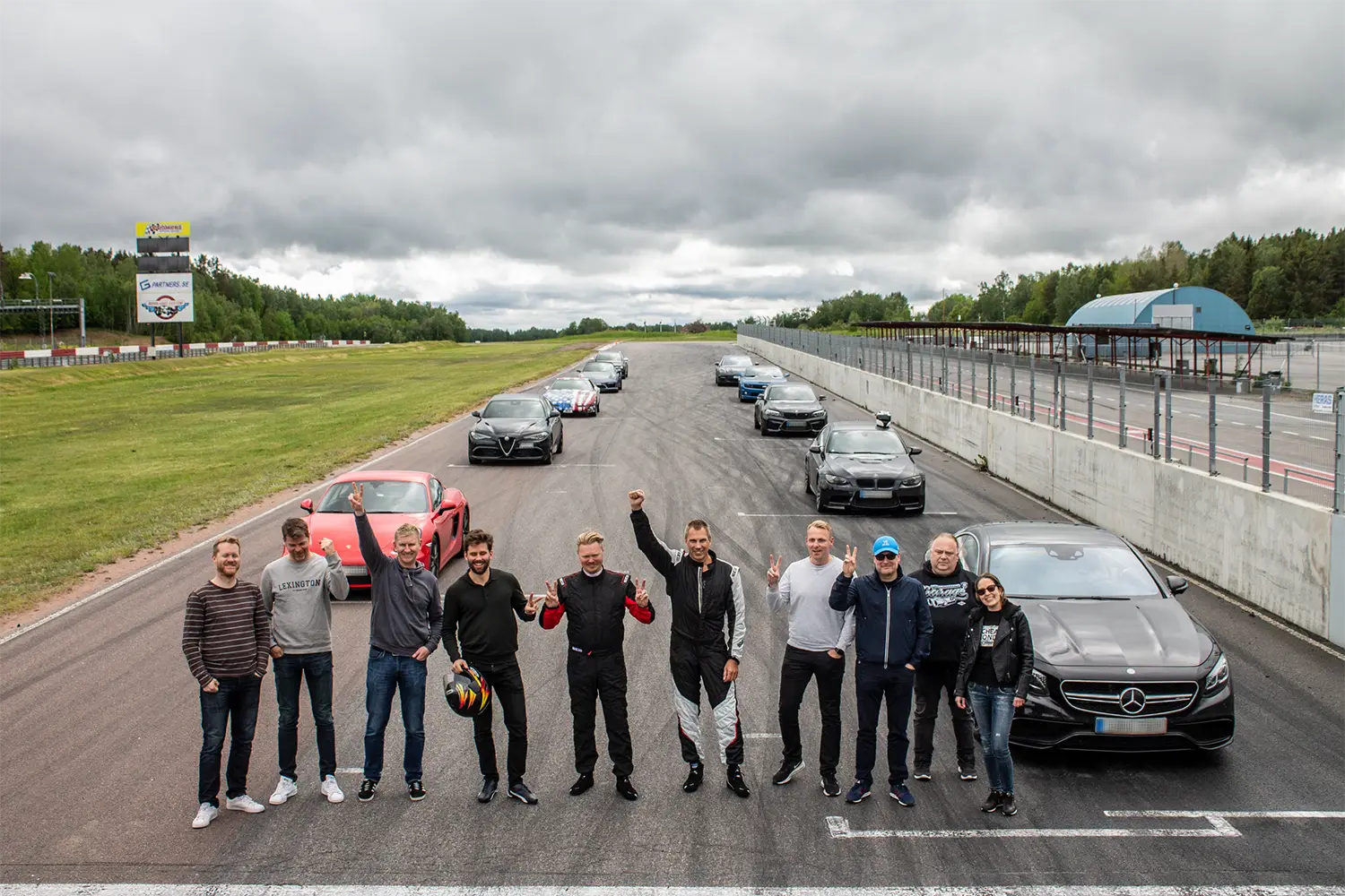 En grupp människor, några i racingutrustning, står och ler på en racerbana med en rad sportbilar bakom sig. Himlen är mulen, och det finns gräs och träd till vänster och en byggnad till höger.
