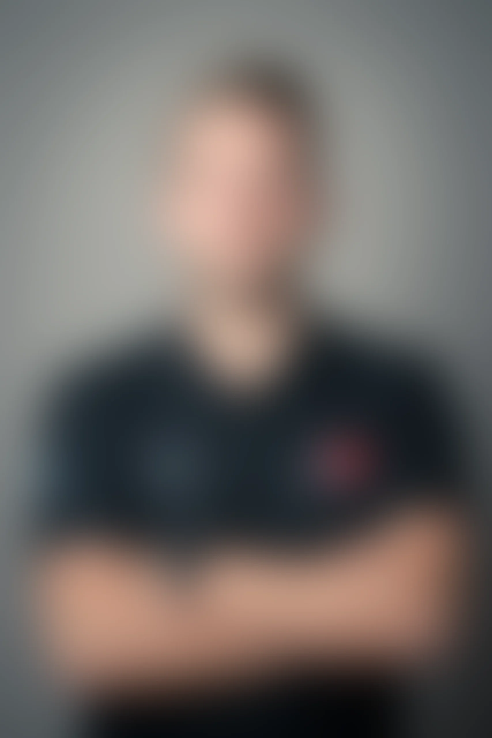 Suddig bild av en person som står mot en grå bakgrund, med armarna i kors. Personen är klädd i en mörk skjorta med otydliga logotyper och ett rött emblem.