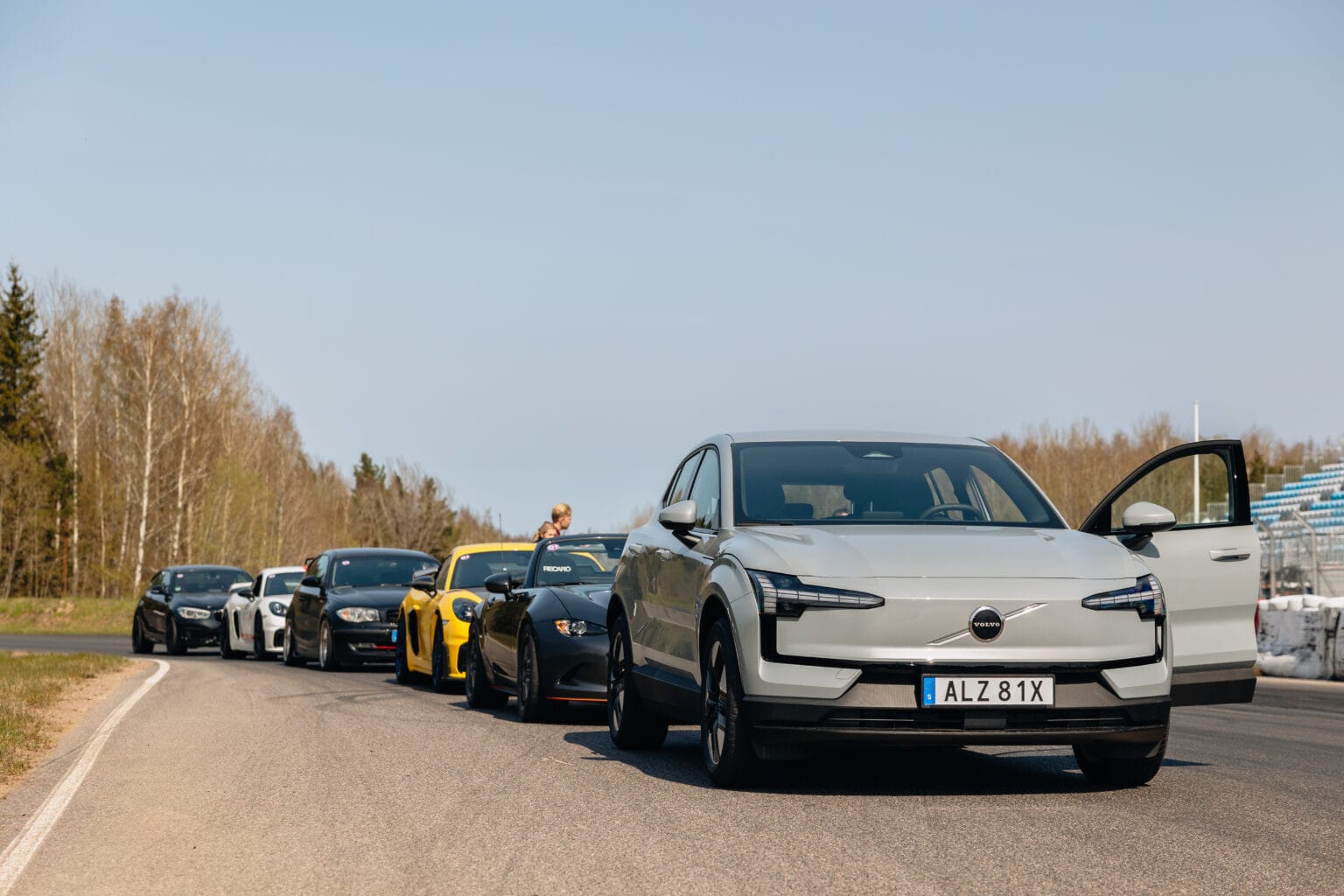 En rad bilar, inklusive en vit Volvo SUV framtill med förardörren öppen, står parkerade på en racerbana. Flera andra sportbilar står uppradade bakom den under en klar himmel.