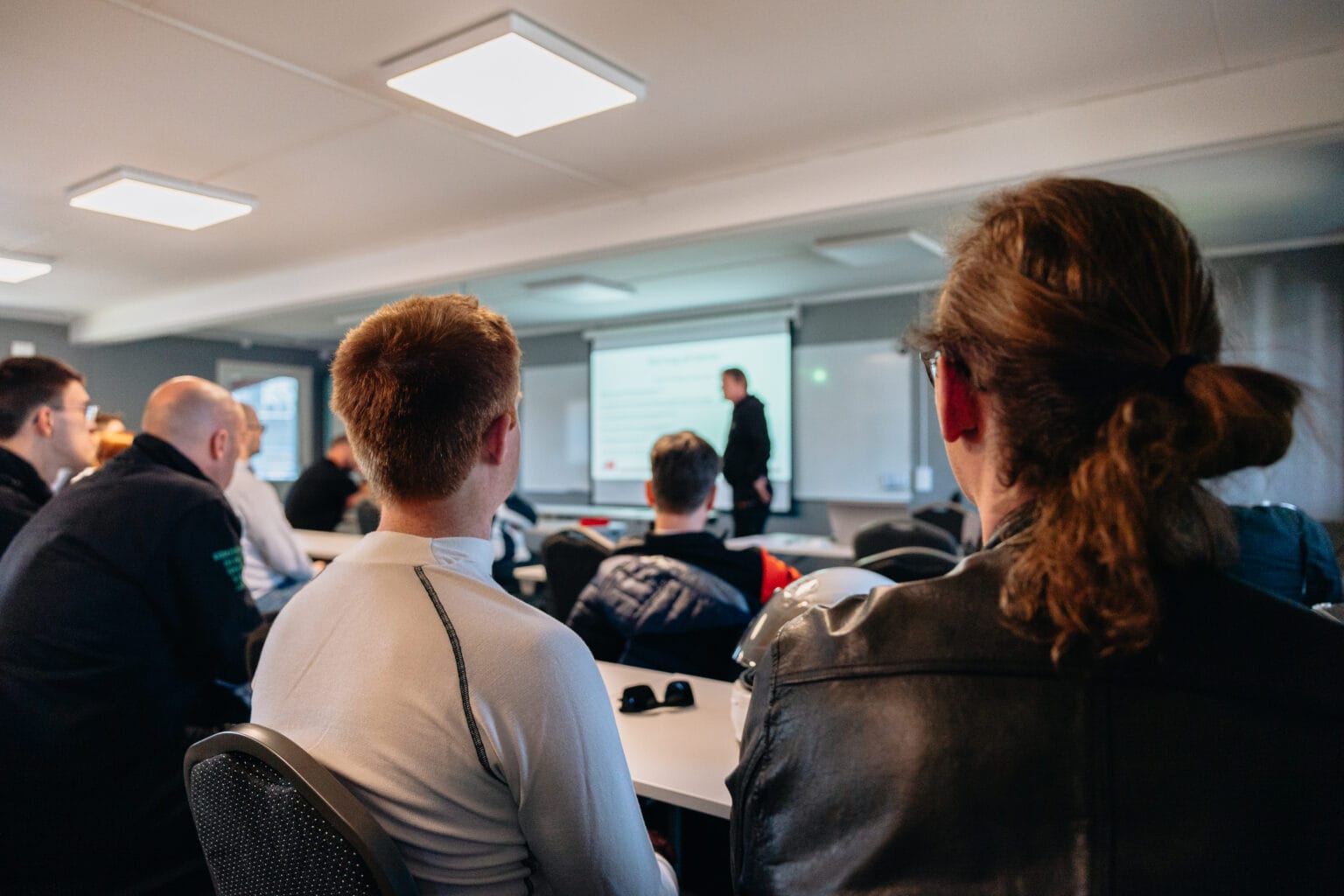 En grupp människor sitter i ett klassrum mitt emot en presentatör som står nära en projektionsskärm med en presentation uppvisad. Fokus är på publiken, med presentatören suddig i bakgrunden.