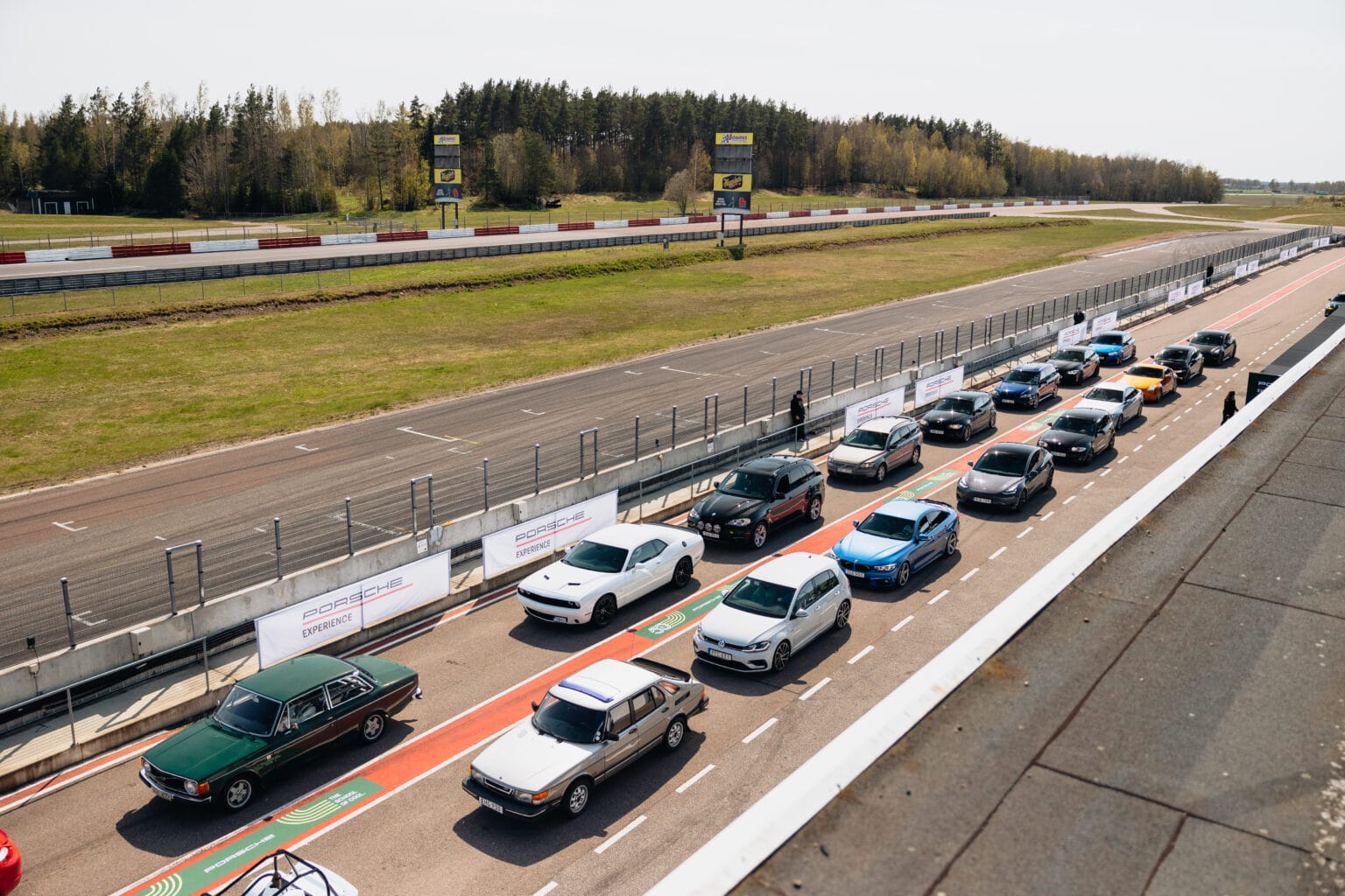 Bilar av olika märken och färger står uppställda sida vid sida i två rader på en depåbana. Banan är avgränsad av staket och träd syns i bakgrunden under en klar himmel.