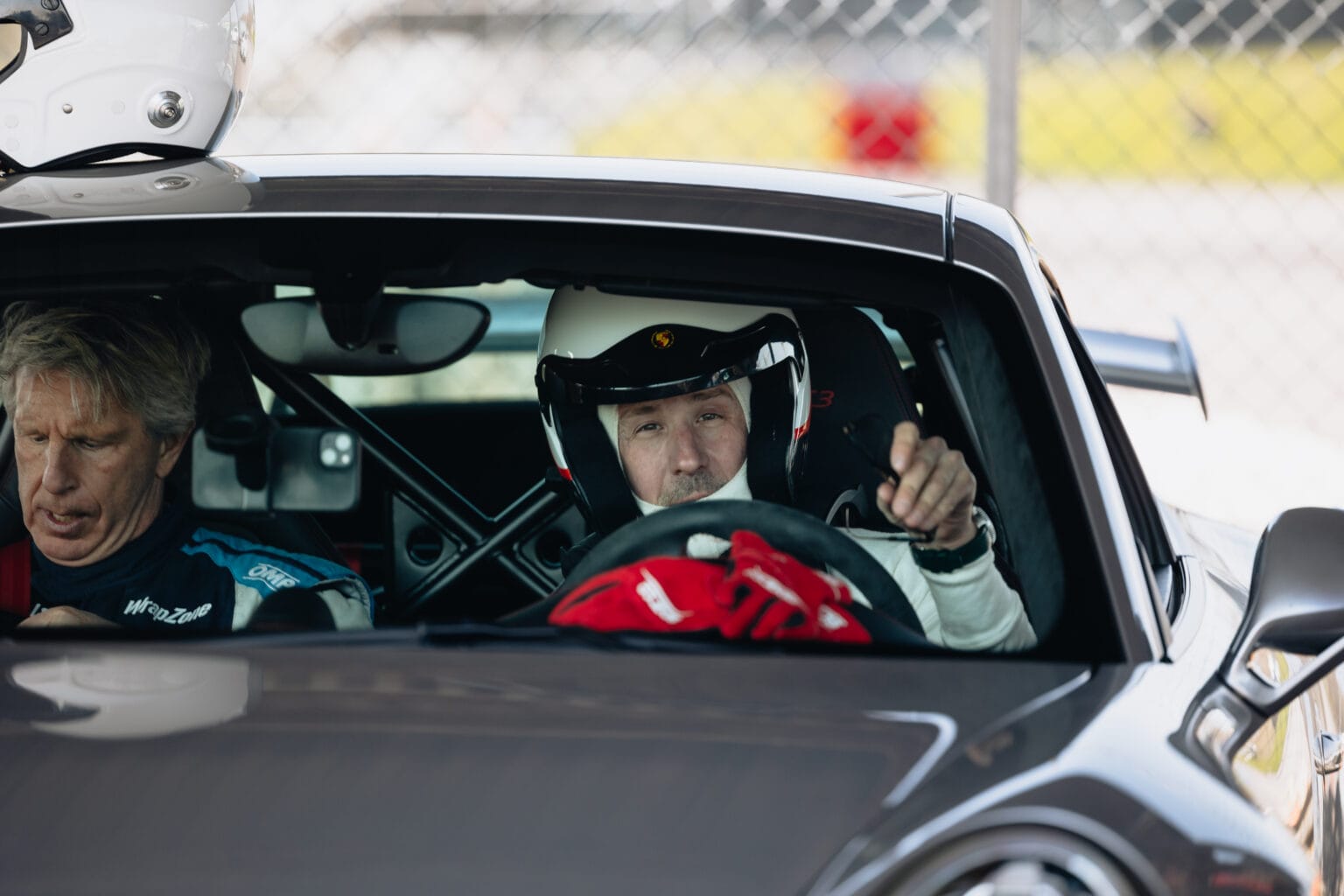 Två män sitter i en sportbil; föraren bär en racerhjälm och pekar framåt, medan passageraren tittar ner på något i sina händer. En racersele och en säkerhetsbur syns inuti bilen.