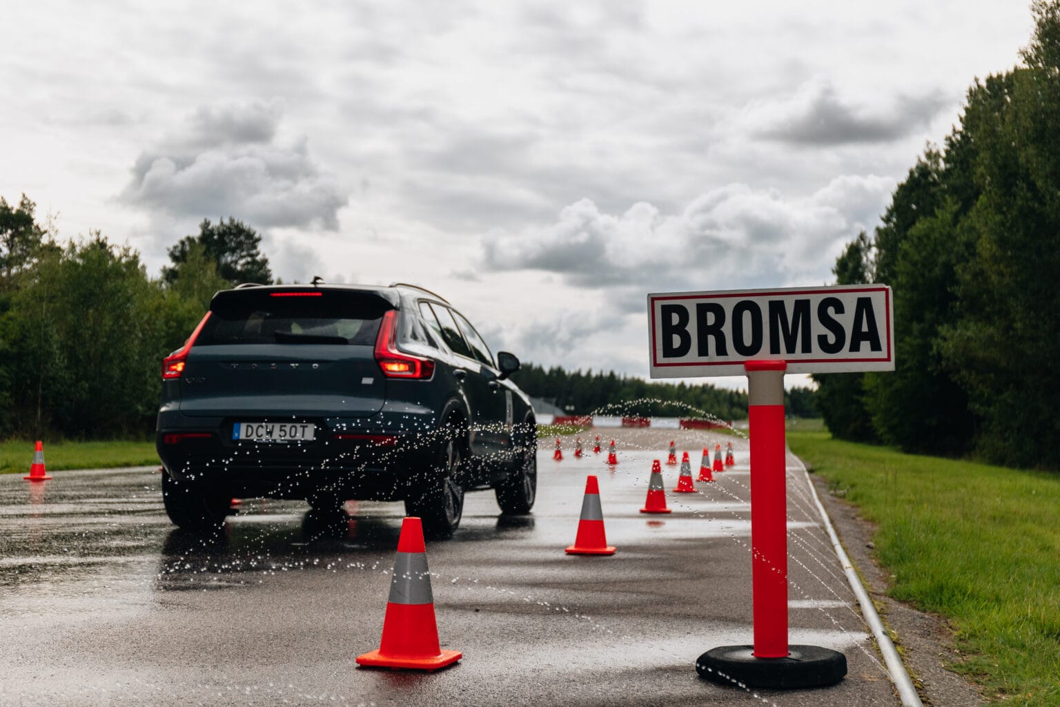 En bil som kör på en våt väg kantad med orange koner och passerar en skylt med texten BROMSA. Vatten stänker från däcken och landskapet är omgivet av träd under en molnig himmel.