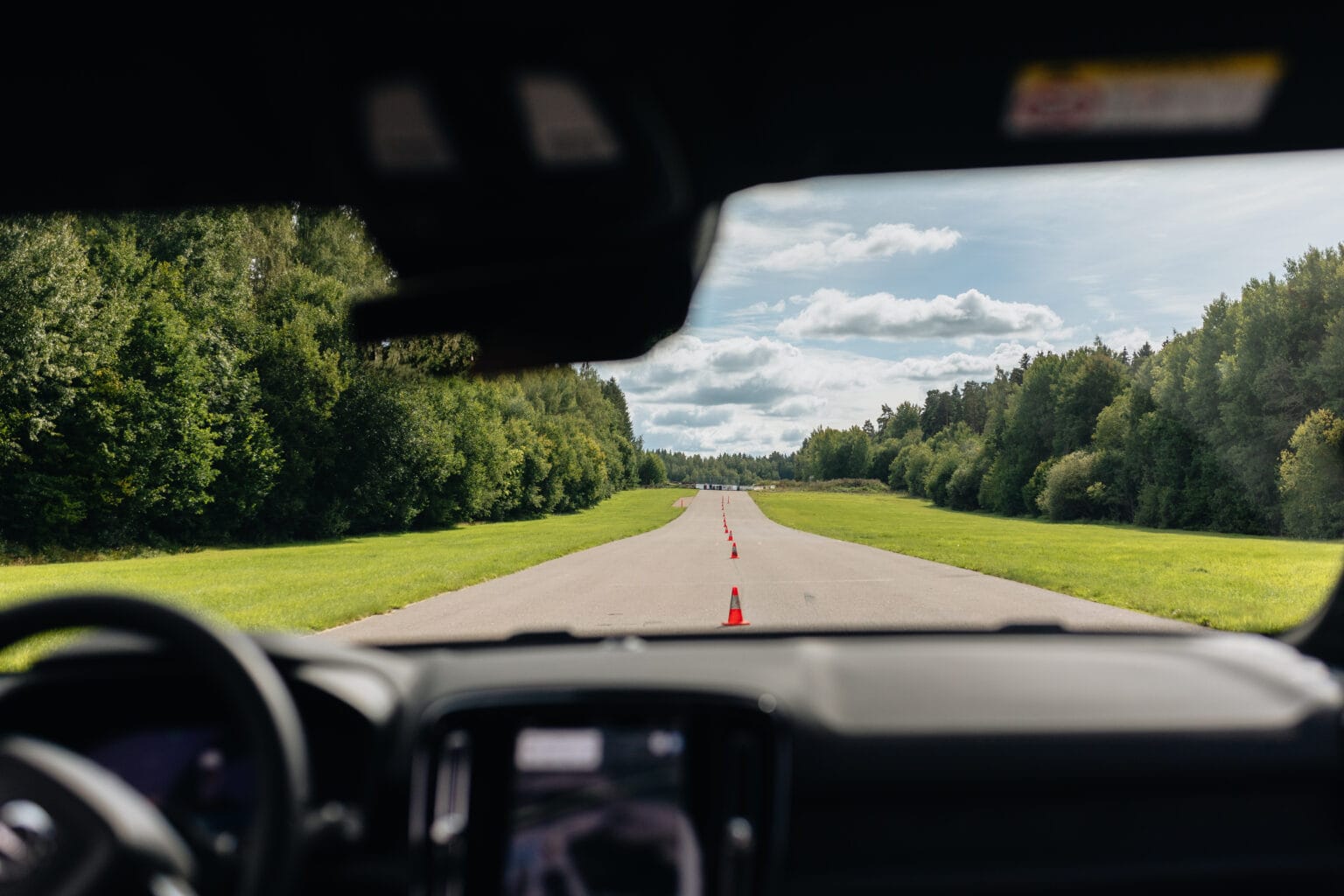 Vy inifrån en bil som tittar ut genom vindrutan på en tom väg kantad av orange trafikkoner, omgiven av gröna träd under en delvis molnig himmel.