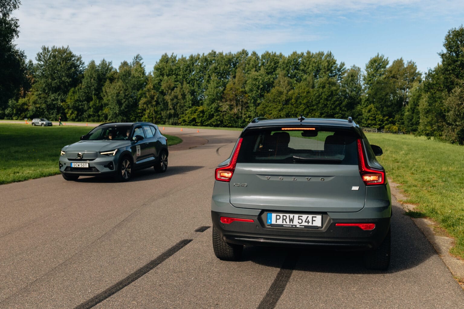 Två grå Volvo-SUV:ar på en asfalterad väg, en i förgrunden sedd bakifrån och den andra i bakgrunden närmar sig från motsatt håll, omgivna av grönt gräs och träd under en blå himmel.