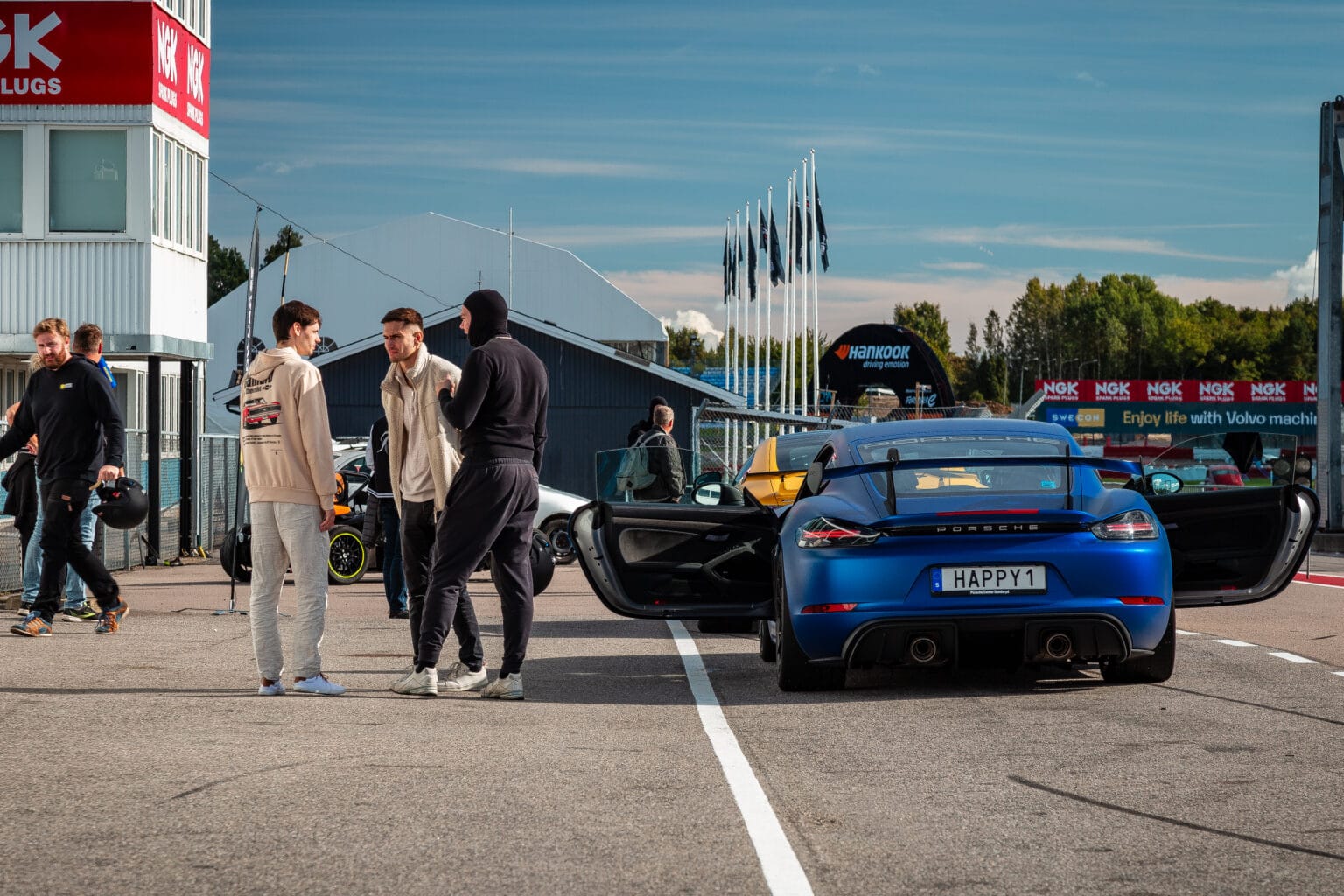 En blå Porsche med öppna dörrar står parkerad på en racerbana. Tre män står och pratar i närheten, medan andra och diverse bilar syns i bakgrunden. Flaggor och byggnader kantar banan under en delvis molnig himmel.