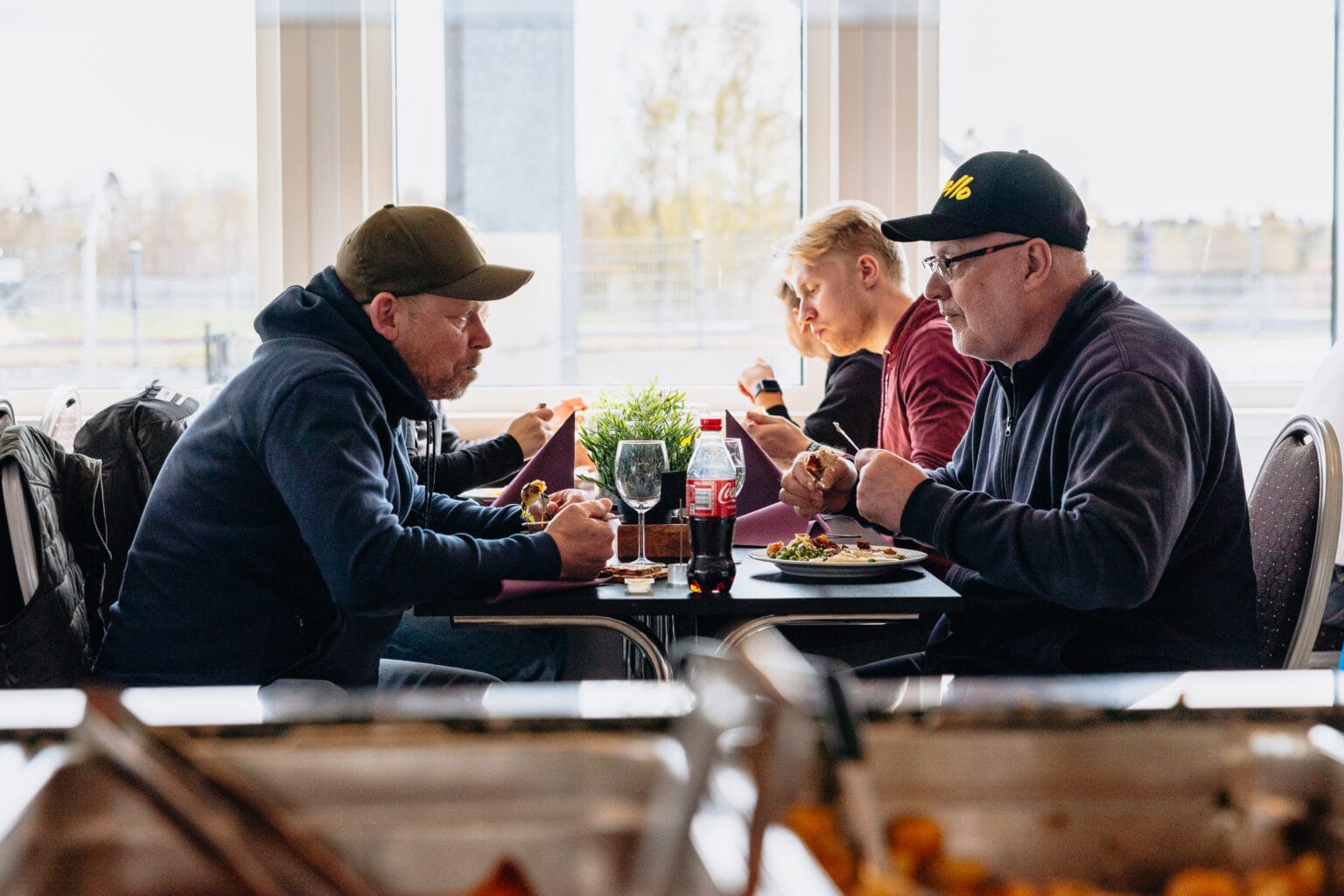 Fyra män sitter vid ett restaurangbord och äter en måltid tillsammans. Tallrikar med mat, drycker och en krukväxt står på bordet. Stora fönster i bakgrunden släpper in dagsljus. Bilden är inramad av brickor med mat i förgrunden.