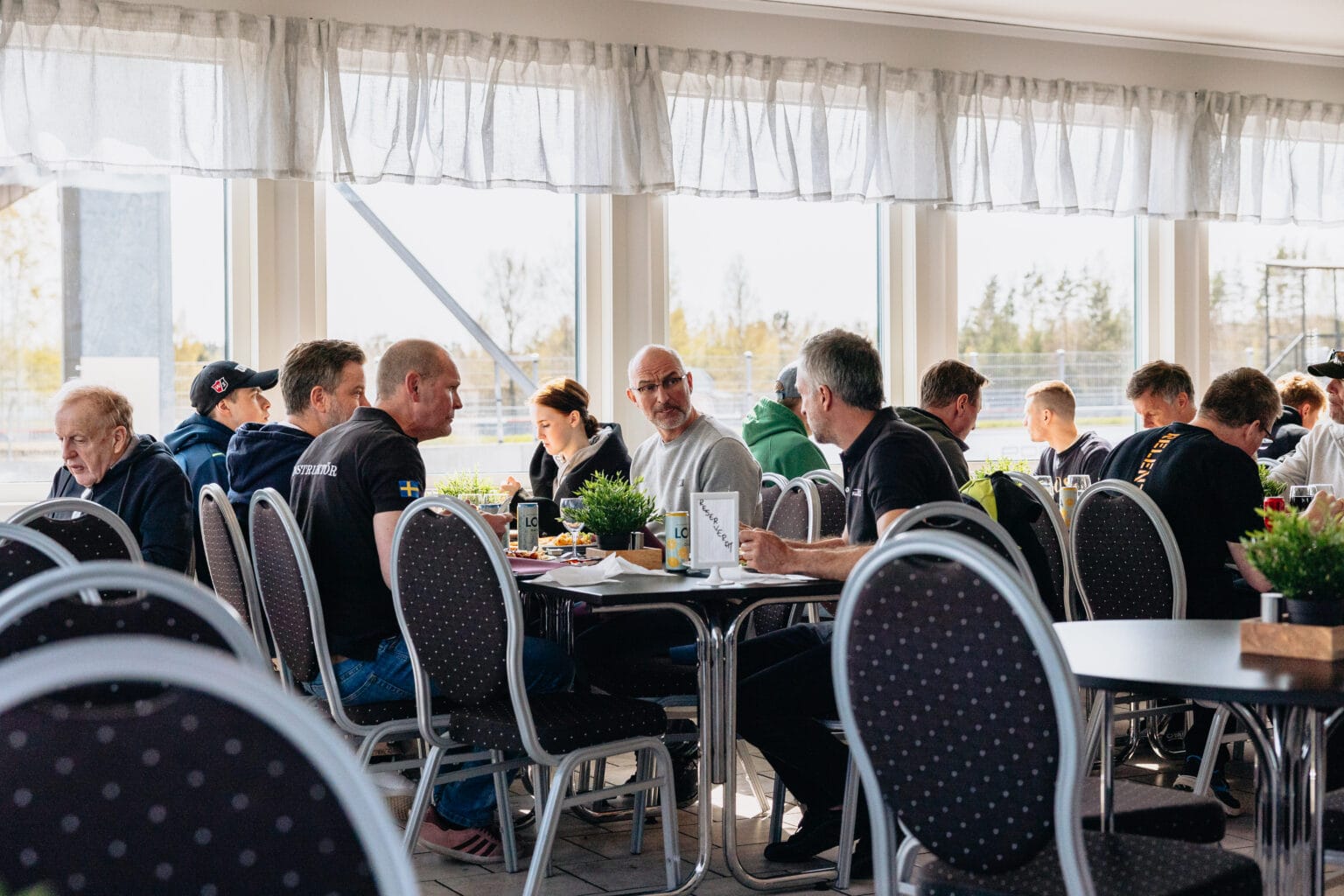 En grupp människor sitter vid bord i en ljus, solbelyst matsal och äter och pratar. Rummet har stora fönster, stolar av metall och krukväxter på borden.