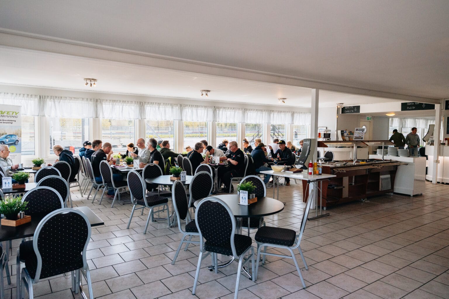 En grupp människor sitter vid bord i en ljus och rymlig cafeteria med stora fönster, svarta stolar och växter på borden. Några äter, några pratar och andra står nära matdisken.