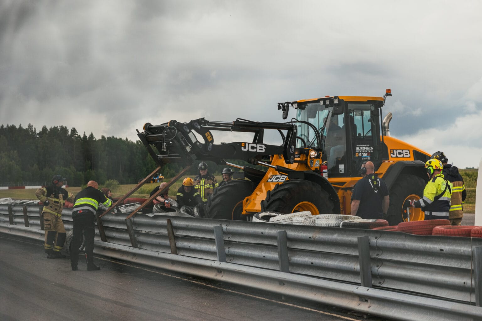 Brandmän och räddningspersonal använder ett JCB-byggfordon för att ta bort skräp från en metallbarriär på en racerbana, under molnig himmel, med träd i bakgrunden.