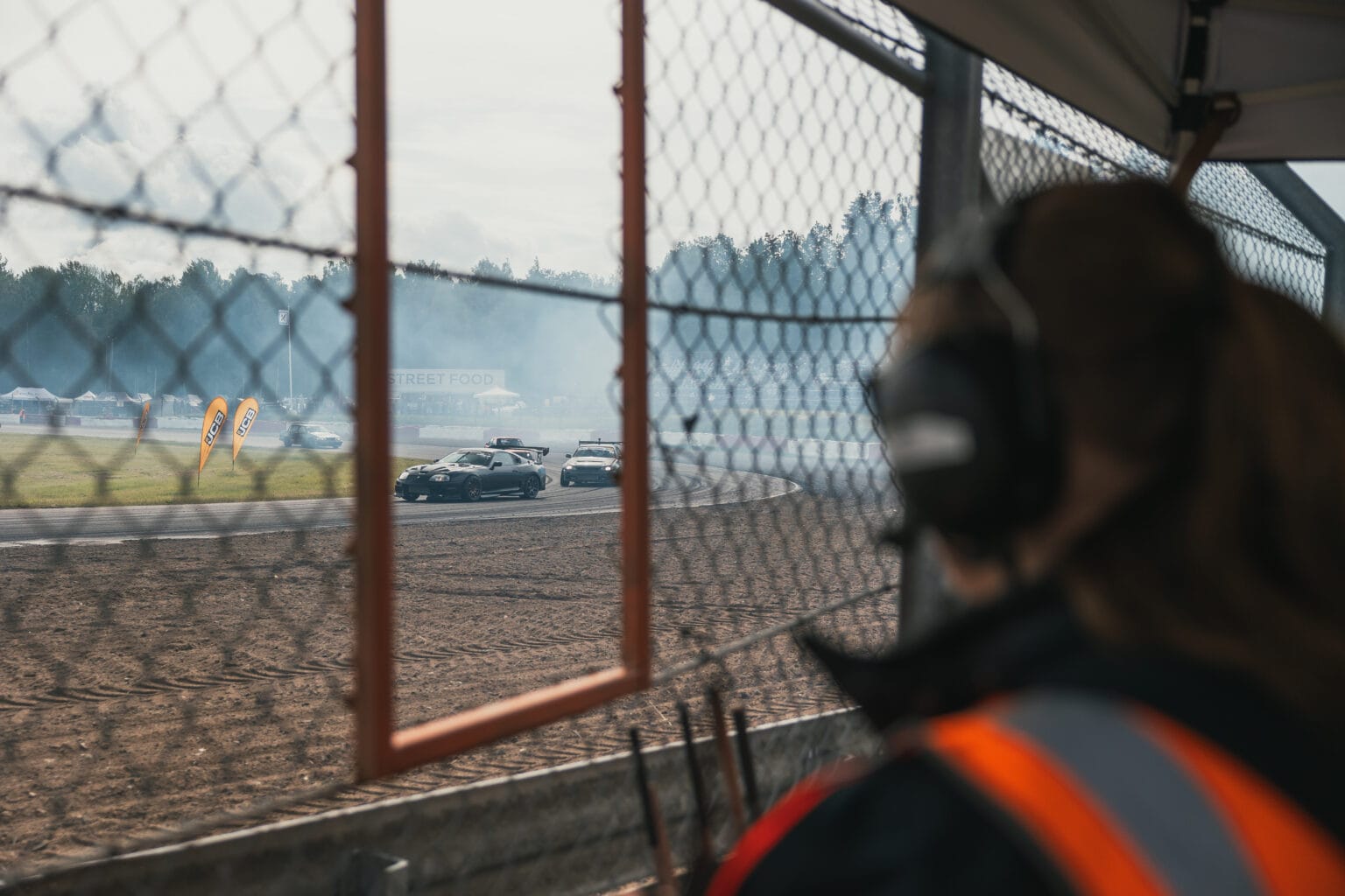 En person med headset och skyddsväst tittar på bilar som driver runt på en racerbana, som ses genom ett kedjelänkstaket med däckrök och träd i bakgrunden.
