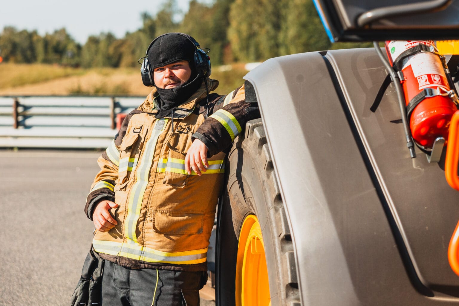 En brandman iförd skyddsutrustning och hörselkåpor lutar sig mot ett stort gult däck från ett byggfordon utomhus en solig dag, med träd och ett räcke i bakgrunden.