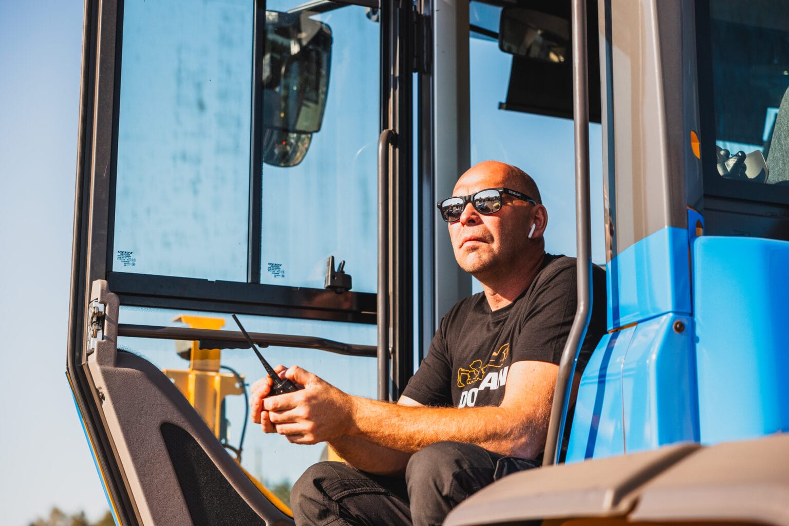 En man i solglasögon och svart t-shirt sitter i den öppna dörröppningen på en blå byggbil, håller i en walkie-talkie och tittar tankfullt ut i fjärran.