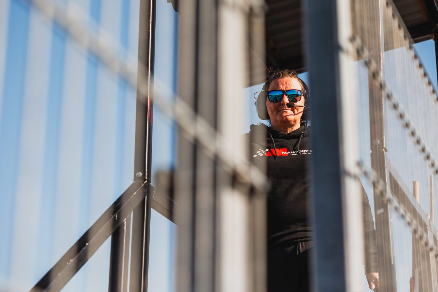 En person i solglasögon och svart huvtröja står på en metalltrappa, där solljus och metallstänger skapar skuggor och reflektioner runt dem. Himlen är klar och blå i bakgrunden.