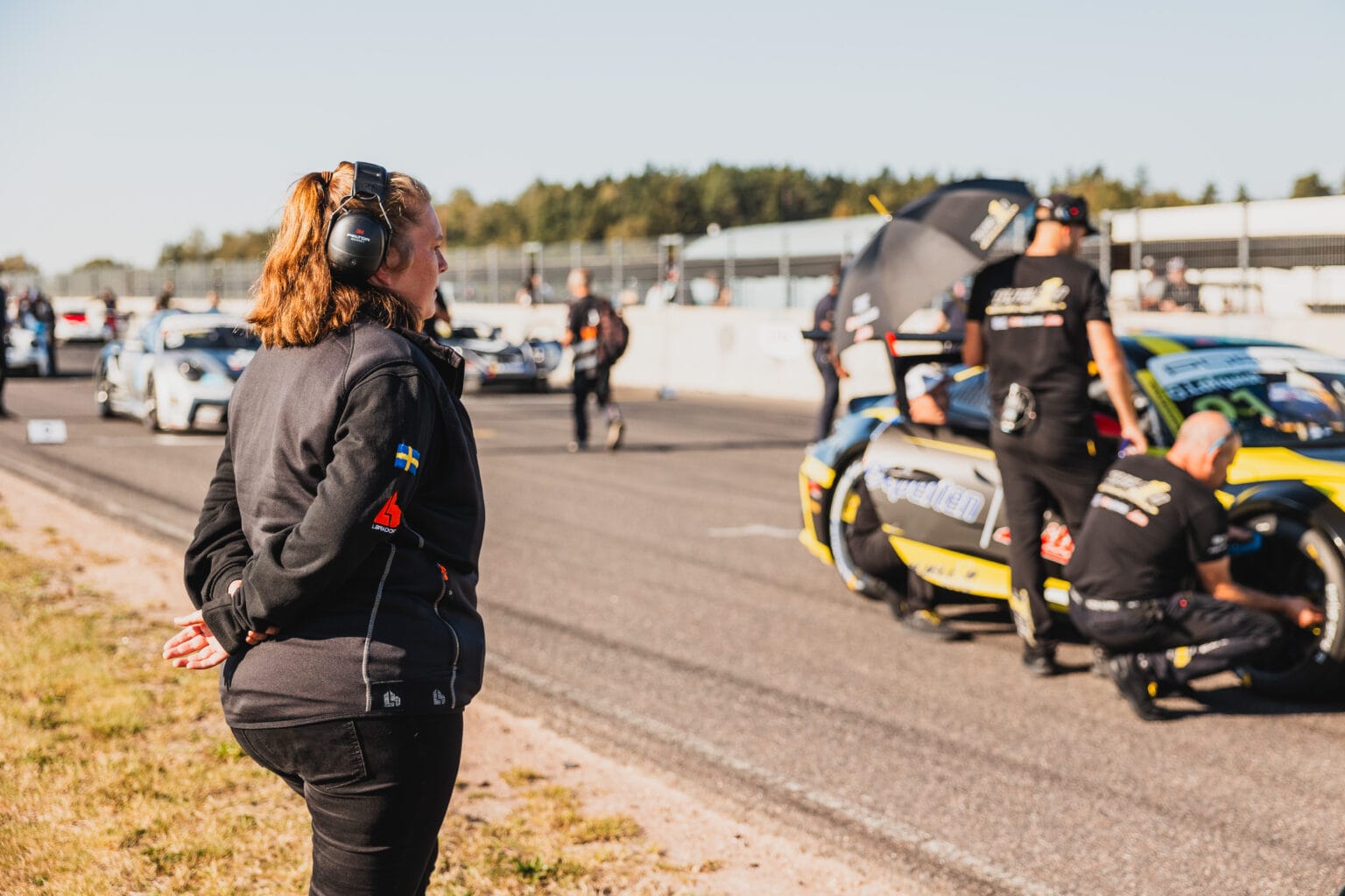 En kvinna med hörlurar och en jacka med en svensk flagga på ärmen står vid sidan av en racerbana och tittar på när personalen arbetar med racerbilar som är uppställda för ett evenemang en solig dag.