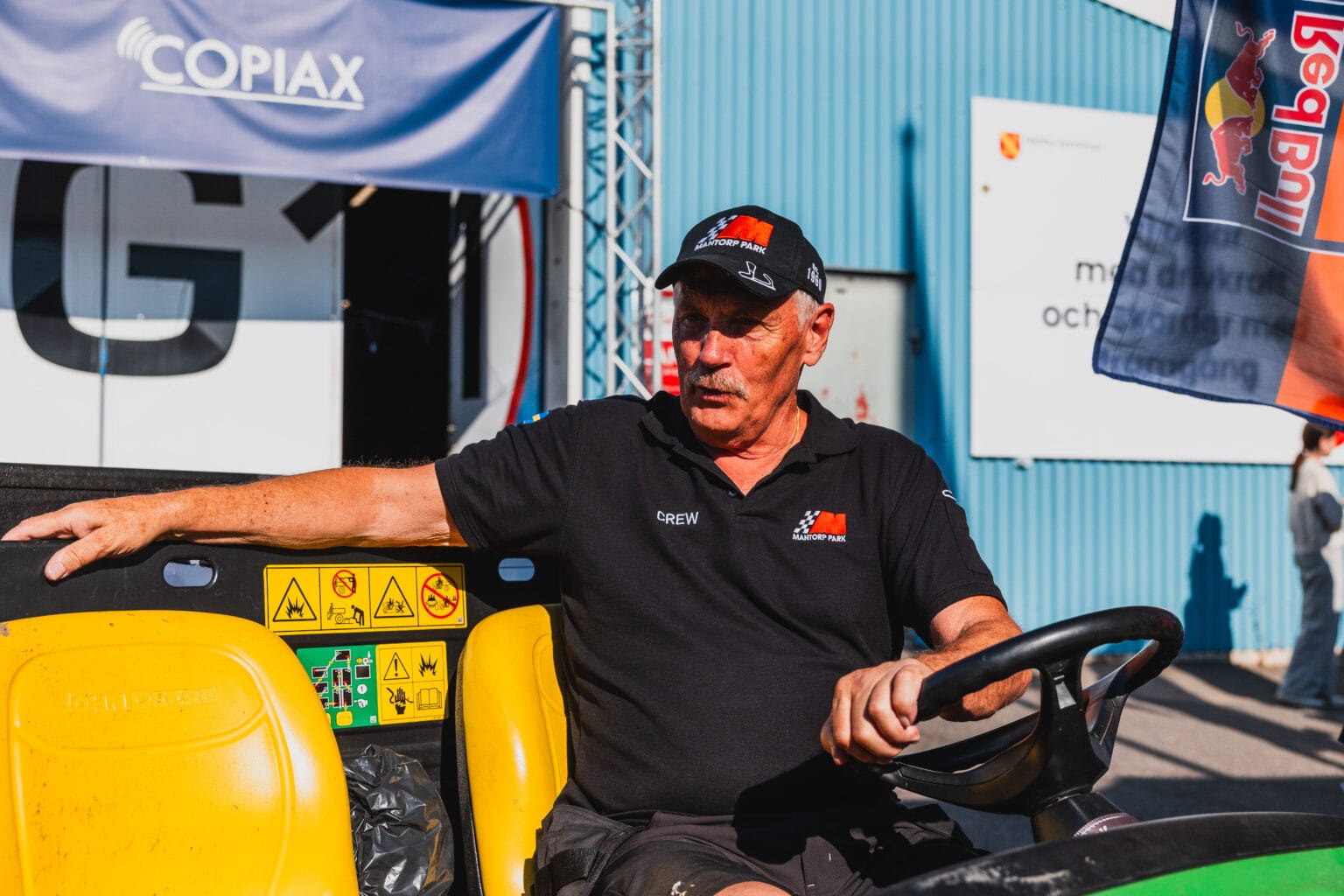 En äldre man med svart keps och polotröja sitter i förarsätet på en golfbil och pratar med ena armen vilande på ryggstödet, mot en utomhusbakgrund med banderoller och en blå byggnad.