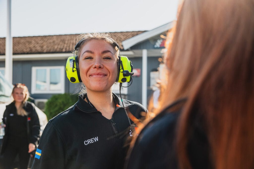 En kvinna med headset och jacka med CREW-märkning ler mot kameran utomhus, med en annan person i förgrunden och en byggnad i bakgrunden en solig dag.