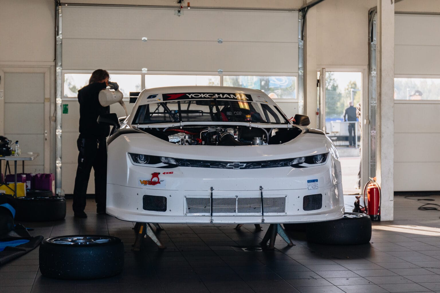 En vit racerbil med öppen motorhuv står i ett garage medan en mekaniker arbetar med motorn. Reservdäck och utrustning finns runt omkring och solljuset strömmar in genom öppna dörrar i bakgrunden.