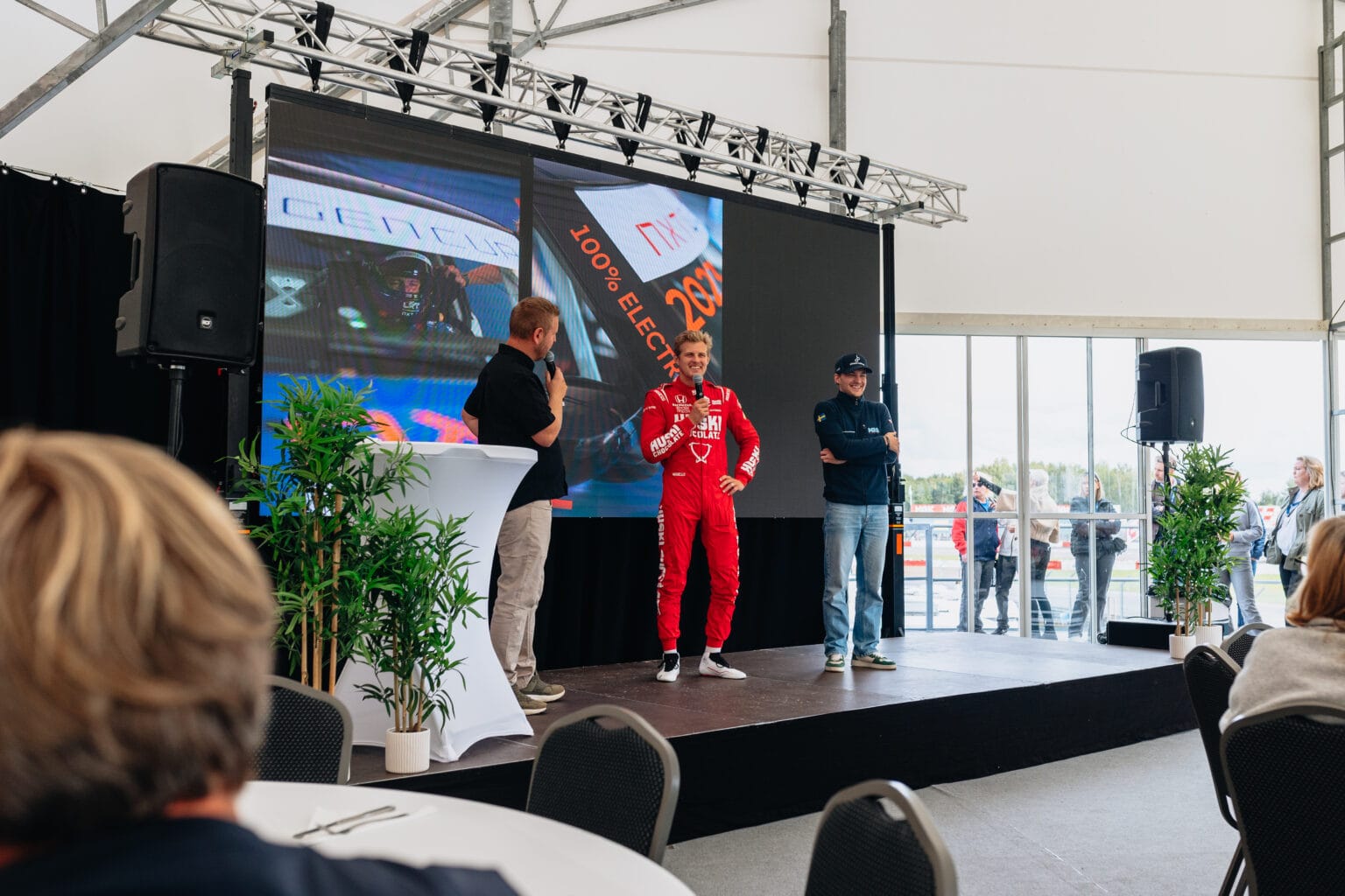 En person i röd racingdräkt står på scenen mellan två personer som håller i mikrofoner, framför en stor skärm och en publik på ett inomhusevenemang med växter och bord.