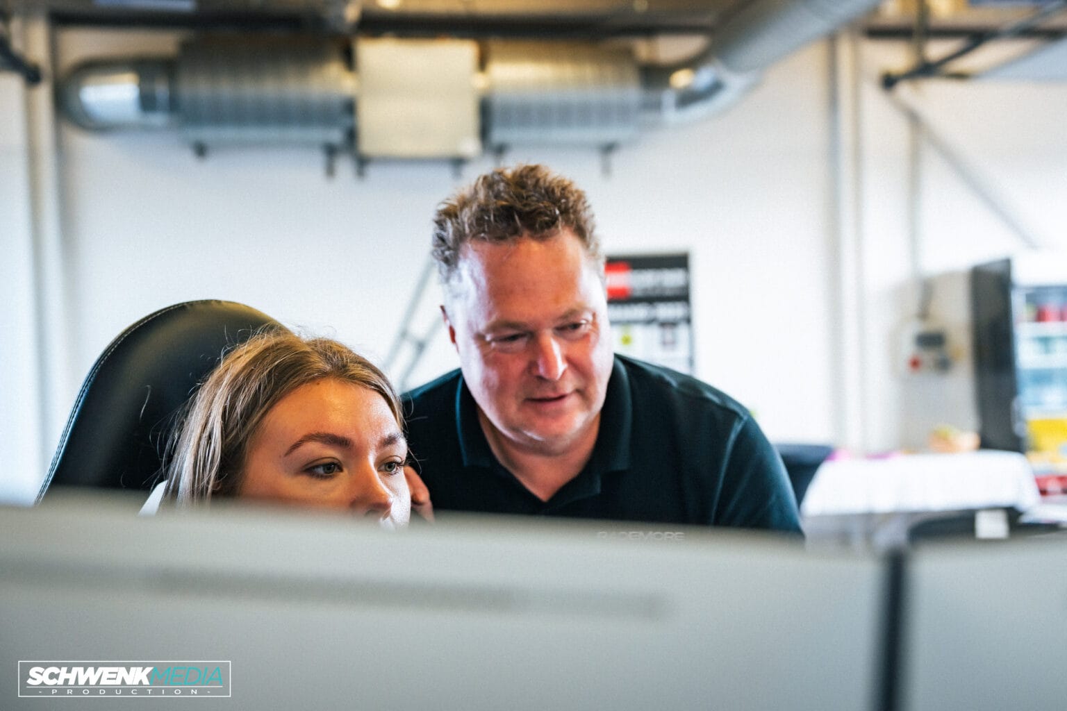 En man och en kvinna tittar på datorskärmar i ett kontor med industriella ventilationskanaler i bakgrunden. Mannen står bredvid kvinnan, som sitter ner, medan de fokuserar på bildskärmarna.