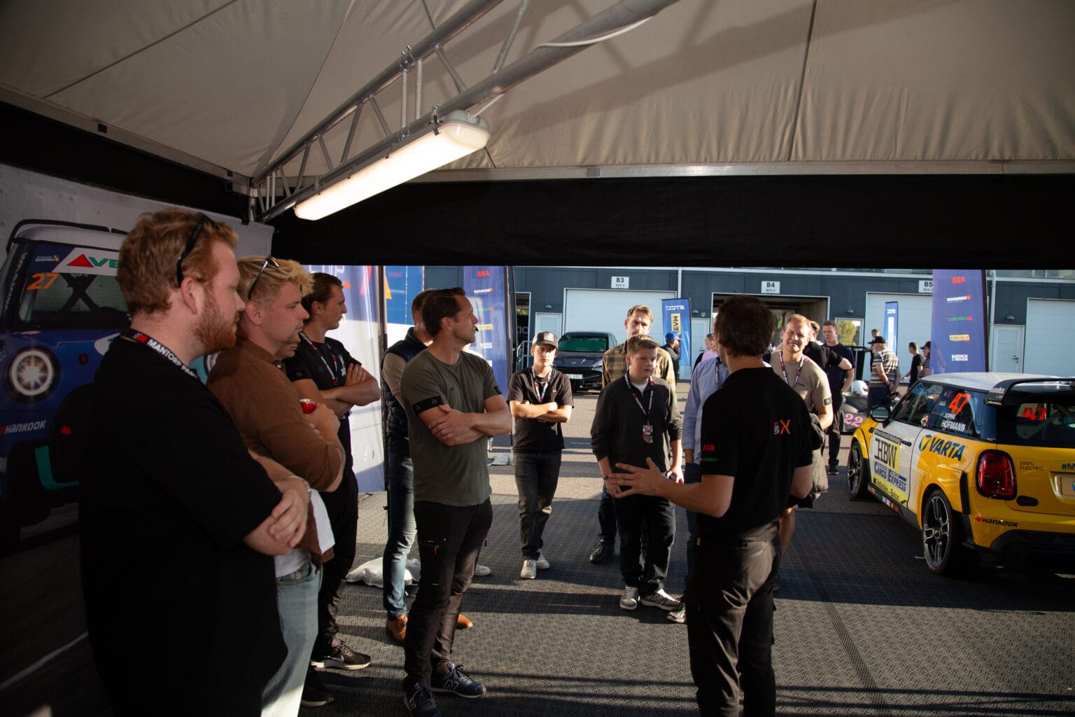 En grupp människor står i ett tältområde bredvid två racerbilar och lyssnar på en person som talar och gestikulerar med sina händer. Solljuset flödar in och kastar skuggor på golvet.