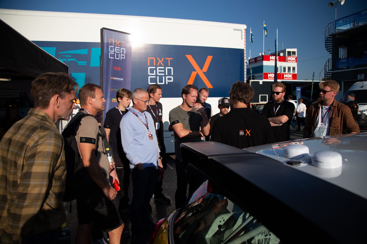 En grupp människor står i solljus och lyssnar på en person som talar bredvid en racerbil. En trailer med texten NXT GEN CUP är parkerad i närheten. Flaggor och eventbås syns i bakgrunden.