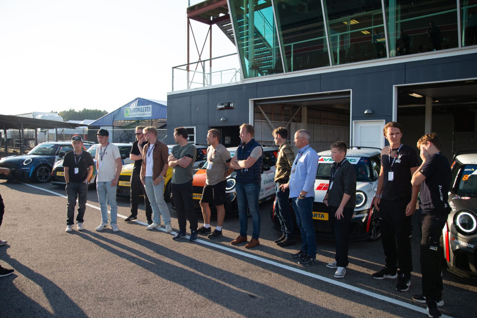 En grupp människor står i en rad utomhus bredvid racerbilar och en byggnad med stora fönster, som kastar långa skuggor på marken i solljuset.